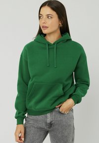 Sweat à capuche vert en matériau doux, avec une poche kangourou, des cordons ajustables et des poignets côtelés.