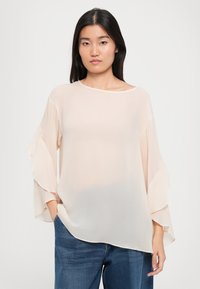 Genomskinlig beige blus med breda, lager-på-lager ärmar och rundad halsringning, tillsammans med blå denimjeans. Slät textur och lätt material.