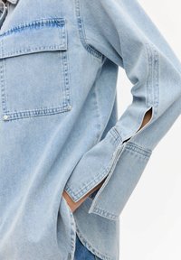 Camicia in denim azzurro chiaro con due tasche sul petto, dettagli con bottoni a pressione e spacchi laterali sui polsini. Presenta una vestibilità comoda e una consistenza morbida.