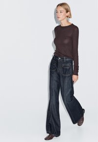 Dark brown long-sleeve top paired with wide-leg dark denim jeans featuring front pockets and white stitching, complemented by brown heeled shoes.