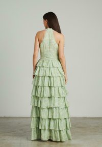 Zielona, kwiecista sukienka maxi z halterem, dopasowanym korpusem i warstwowym, falbankowym wykończeniem. Materiał wydaje się lekki i teksturowany.