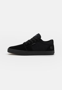 Black sneaker with a suede and canvas upper, round toe, flat rubber sole, and tonal laces. Features a small logo on the side.