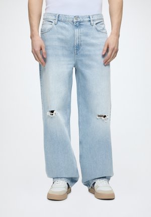 Ανοιχτό μπλε τζιν με φαρδιά μπατζάκια και σκισίματα στα γόνατα, συνδυασμένα με λευκά και μπεζ αθλητικά παπούτσια και λευκό τοπ.