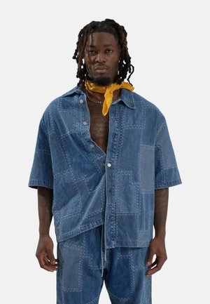 Hombre con rastas que lleva una camisa y pantalones de mezclilla patchwork, bandana amarilla alrededor del cuello, de pie frente a un fondo blanco liso.