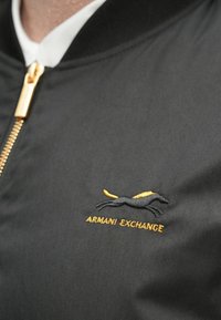 Μαύρο bomber μπουφάν με χρυστό φερμουάρ · διαθέτει μαύρο και χρυσό κεντημένο λογότυπο. Λείο ύφασμα με υφασμένη υφή.