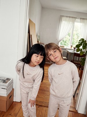 Deux enfants portant des tenues Nike claires assorties se penchent vers l’appareil photo dans une pièce lumineuse avec un parquet et de grandes fenêtres.