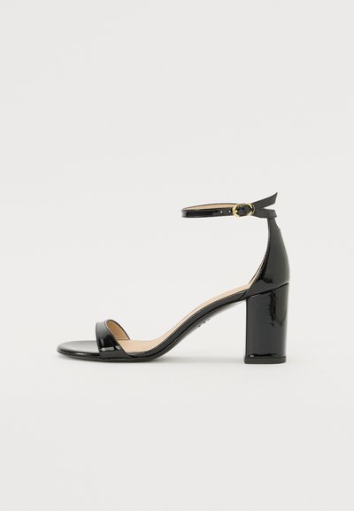 Sandale à talon haut en cuir verni noir avec bride à la cheville et boucle dorée, bout ouvert et talon bloc sur fond blanc.