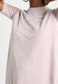 Przesadna różowa koszulka z miękką fakturą, z subtelnym wytłaczanym logo Adidas i akcentami w kolorze szarym przy kołnierzyku.