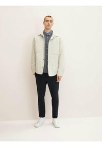 Chaqueta ligera de color beige claro con dos bolsillos en el pecho, llevada sobre una camisa azul oscura, combinada con pantalones negros y zapatillas blancas.
