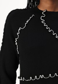 Svart ribbestrikket genser med dekorative hvite volanger langs skuldrene og kanten. Tettsittende design med teksturert stoff.