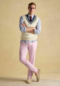 Kremowa dzianinowa kamizelka z wzorem warkocza, granatowo-białe paski przy dekolcie, noszona na paskowanej koszuli. Jasnoróżowe spodnie i beżowe mokasyny.
