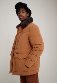 Casaco acolchoado castanho com gola escura peluda, apresenta botões de pressão e dois bolsos frontais. Desenhado com um corte descontraído e textura suave.