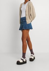 Beige knit cardigan with large buttons, white tank top, denim pleated skirt, and chunky black sandals with white fur accents. Purple glitter socks.