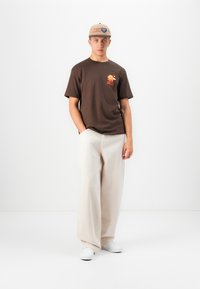 Ung mand iført en beige kasket, brun T-shirt med orange grafik, brede cremefarvede bukser og hvide sneakers, stående foran en ensfarvet hvid baggrund.