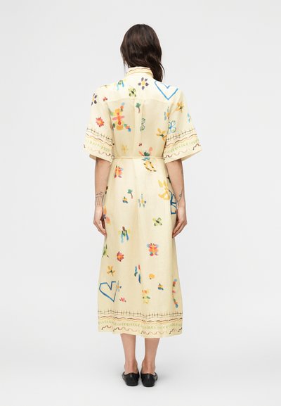 Γυναίκα όρθια με την πλάτη προς τα εμπρός, φοράει κρεμ μίντι φόρεμα με πολύχρωμα αφηρημένα λουλουδένια και καρδιές σχέδια, συνδυασμένο με μαύρα παντόφλες.