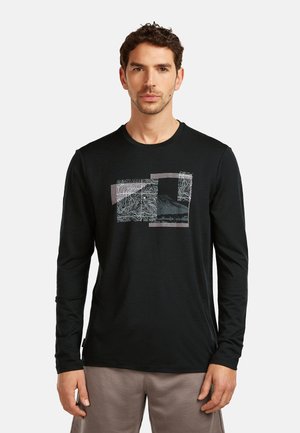 Mężczyzna ubrany w czarną bluzkę z długim rękawem z abstrakcyjnym wzorem topograficznym, stojący na tle jasnego, jednolitego tła, zwrócony przodem.