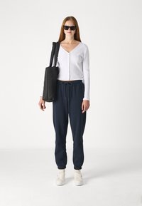 Biely rebrovaný top s dlhým rukávom a gombíkmi, námornícke joggersy a biele tenisky. Čierna taška na rameno a slnečné okuliare dotvárajú outfit.