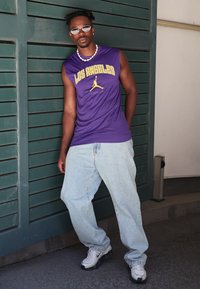 Camisă fără mâneci de culoare purpurie cu textul "LOS ANGELES", blugi largi de un albastru deschis și adidași albi cu accente gri, accesorizată cu ochelari de soare și mărgele.