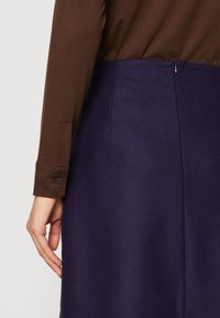 Femme portant un chemisier marron à manches longues rentré dans une jupe taille haute bleu foncé avec une fermeture éclair au dos.