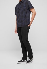 Camicia a maniche corte a quadri nera con accenti blu, due tasche sul petto; abbinata a jeans neri slim-fit e sneaker nere con lacci bianchi.