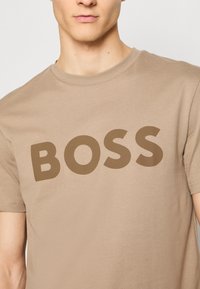 Beige bomulls-T-shirt med en stor, mörkbrun "BOSS"-logga över bröstet. Korta ärmar och klassisk rund halsringning.