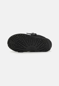 Semelle en caoutchouc noire avec un motif de sculpture texturé, arborant un logo et un accent en boucle au niveau du talon pour faciliter l'enfilage.
