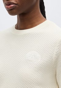 Κοντινό πλάνο ενός ατόμου που φοράει πλεκτό εκρού πουλόβερ με ανάγλυφο λογότυπο EA7 στο στήθος.