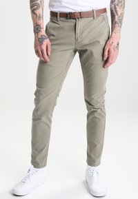 Chinos khaki avec une coupe slim, dotés d'une fermeture éclair, de poches avant et d'une ceinture en cuir marron. Associés à des baskets blanches.