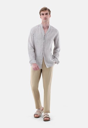 Junger Mann steht und trägt ein hellgraues Leinenhemd mit Knöpfen, beige Hose und beige Sandalen vor einem schlichten weißen Hintergrund.