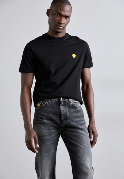 Μαύρο βαμβακερό t-shirt με μικρή κίτρινη γραφική απεικόνιση, συνδυασμένο με ξεθωριασμένα σκούρα γκρι τζιν με χρυσές λεπτομέρειες.