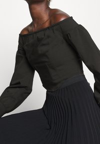 Top nero a spalle scoperte con maniche lunghe a sbuffo, caratterizzato da un corpetto aderente e una texture liscia, abbinato a una gonna scura a pieghe.