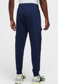 Granatowe spodnie dresowe z elastycznym pasem, kieszeniami bocznymi na cargo oraz ściągaczami przy nogawkach. Połączone z szarymi i neonowozielonymi butami sportowymi.