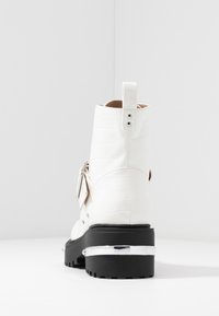 Bottine blanche à texture crocodile, semelle épaisse noire, accent argenté sur le talon, sangle latérale avec boucle et deux boutons à l'arrière.