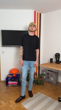 Musthave must-sleeve särgid, helesinised teksad ja pruunid kingad. Seistes toas, kus on televiisor, oranž lamp ja roheline taim.