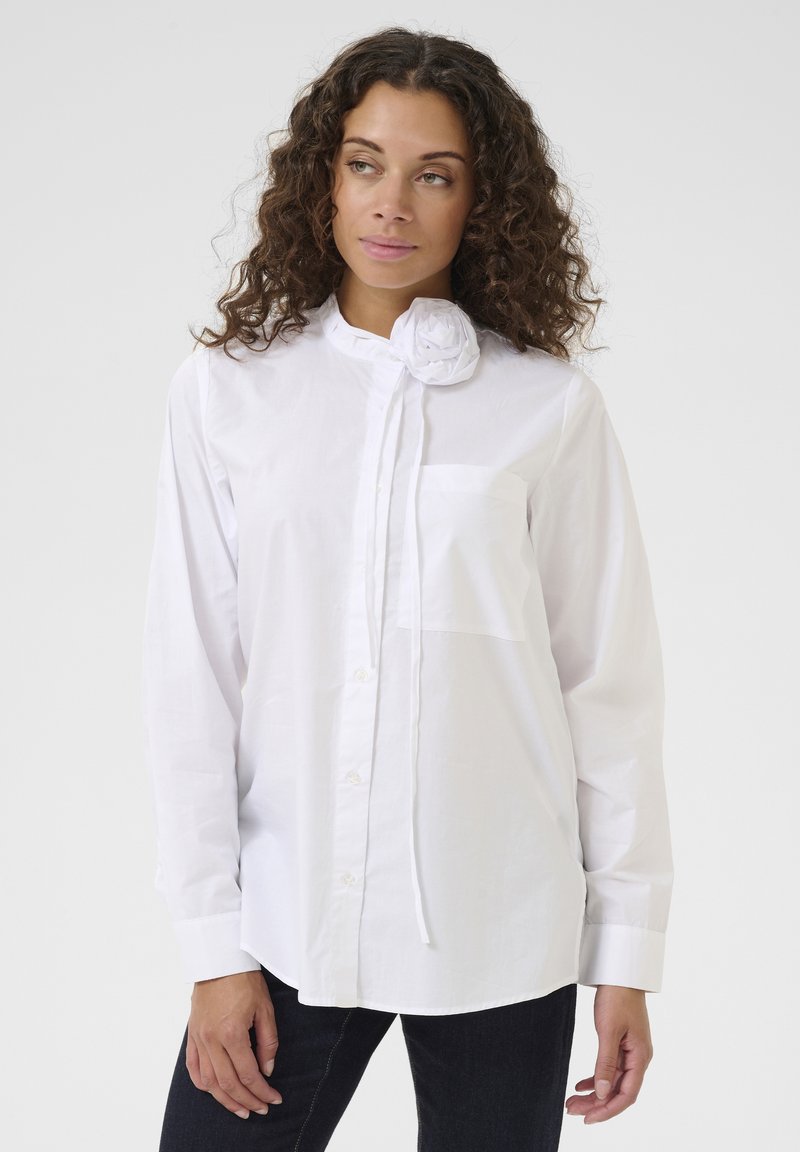 Blusa di cotone bianca con colletto rotondo, bottoni frontali e un accento floreale decorativo al collo. Presenta una tasca sul petto sinistro.