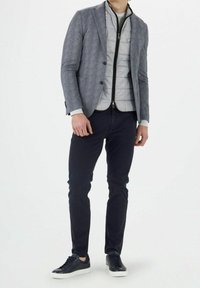 Blazer gris à motif porté sur une veste sans manches matelassée gris clair, associé à un pantalon bleu marine foncé et des baskets noires. Coupe ajustée.