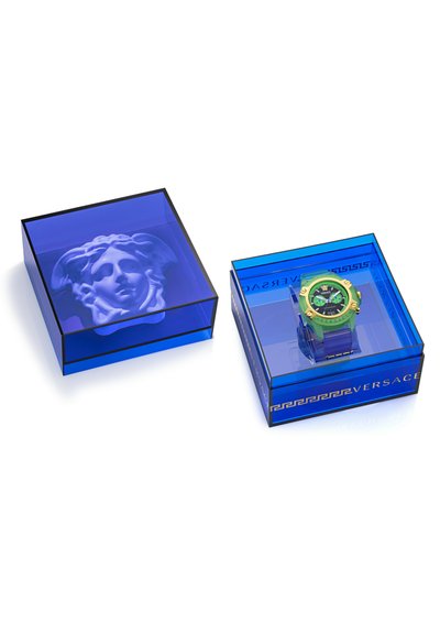 Relógio de pulso verde com mostrador preto numa caixa azul transparente com relevo branco de um rosto. Detalhes em dourado e marca em destaque.