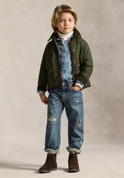 Niño con cabello rubio que está de pie vistiendo una chaqueta verde acolchada, camisa vaquera azul, vaqueros desgastados remangados en los tobillos y botas marrones sobre un fondo neutro.