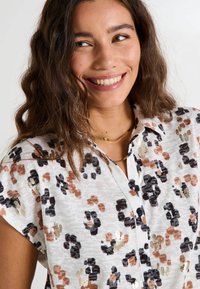 Blouse à manches courtes en couleurs mélangées avec un motif léopard, ornée de accents dorés et en tissu léger texturé. Fermeture à boutons au col.