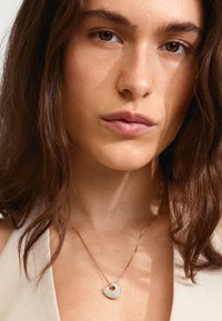 Collier en or avec un pendentif en forme de cœur, orné de pierres précieuses transparentes, porté sur un cou nu sur fond neutre.
