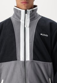 Fleece jacket in black and gray with white striped accents. High collar, full zip, and Columbia logo on the left chest. Soft texture.