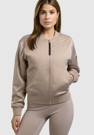 Kobieta w beżowej bomberce na zamek i dopasowanych spodniach, stojąca z jedną ręką w kieszeni na jednolitym tle.