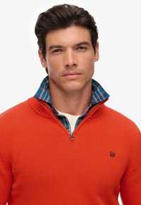 Maglione arancione lavorato a maglia con colletto zip, indossato sopra una camicia a quadri blu e verde. Presenta un piccolo logo sul petto sinistro.