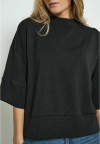 Black, short-sleeve top with a high neckline, featuring a relaxed fit and soft, textured fabric. Part of a casual outfit with jeans.