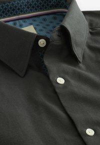 Camisa de botones gris oscuro con una tela texturizada. Presenta un forro de contraste azul con un patrón en el cuello y detalles de rayas multicolores.