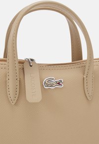 Borsa a tracolla sintetica beige con manici doppi, superficie testurizzata e logo del coccodrillo Lacoste. Presenta una chiusura con zip e un'etichetta con il marchio.