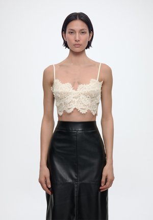 Femeie cu păr negru scurt, purtând un top scurt crem din dantelă cu bretele subțiri și o fustă neagră din piele, cu talie înaltă, stând în fața unui fundal alb.