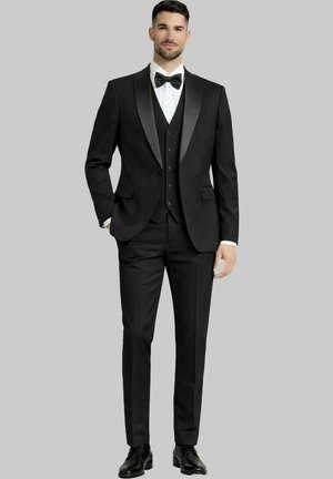 Homme portant un smoking noir à revers en satin, une chemise blanche, un nœud papillon noir, un gilet et des chaussures habillées noires, debout devant un fond gris.