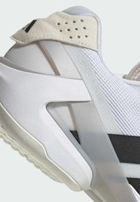 Zapatilla deportiva blanca con parte superior de malla texturizada, rayas negras y un suave acento en beige en el talón. Cuenta con una suela blanca gruesa y un diseño redondeado.