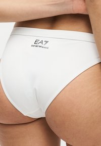 Λευκά μαγιό μπικίνι με ψηλό κόψιμο στο κάτω μέρος της πλάτης και τους γοφούς ενός ατόμου, με μαύρο λογότυπο EA7 Emporio Armani στη μέση της πίσω μέσης.
